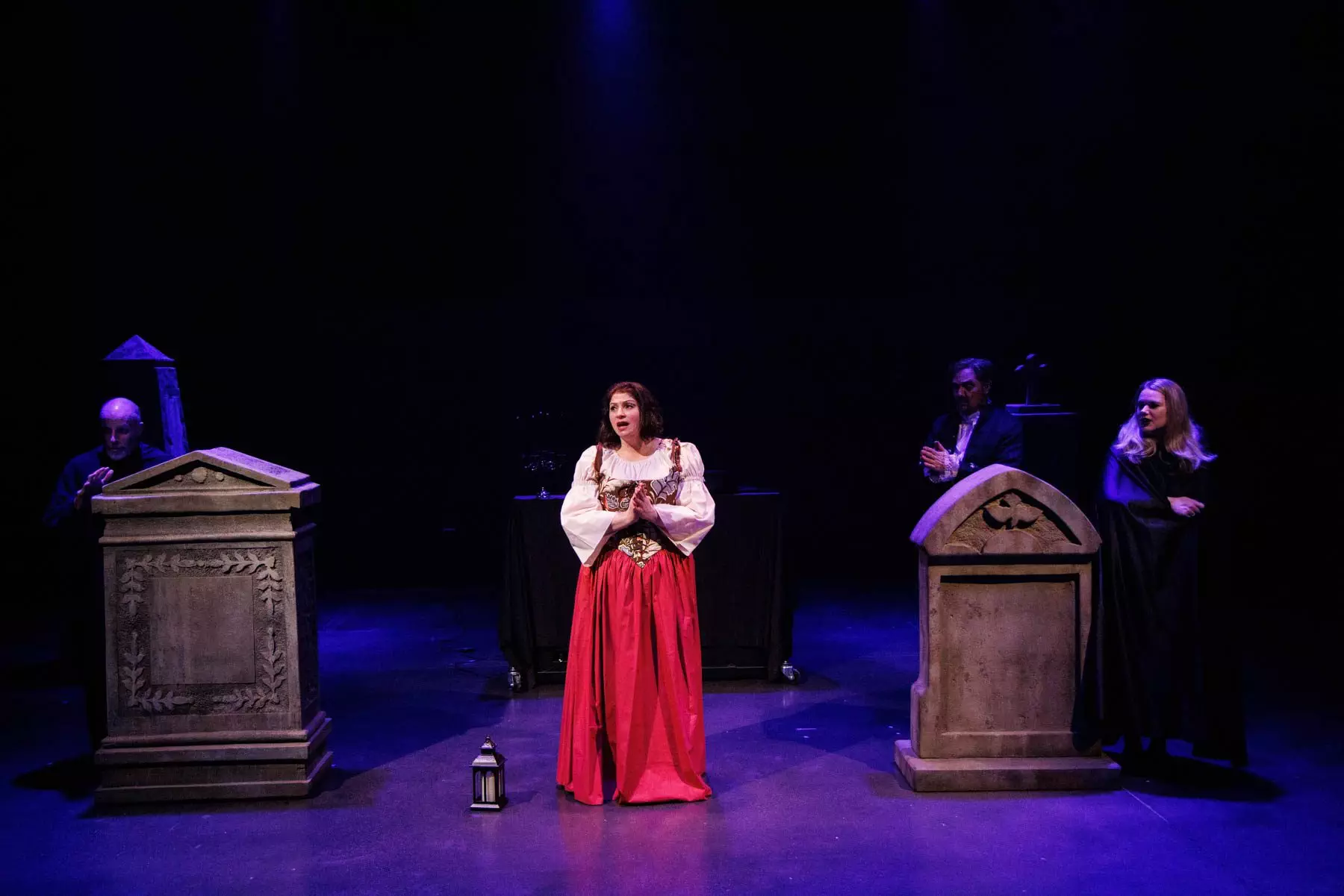Emmy (Yana White) sings "Emmy’s Ballad of the Vampire" from Heinrich Marschner's "Der Vampyr". She wears a beautiful red peasant gown and a white blouse, looking anxious, with a lantern sitting beside her. Among the tombstones surrounding her, other cast members (Doug Brunker, Kyle Hancock, and Holly Howard) lurk in dark clothing.